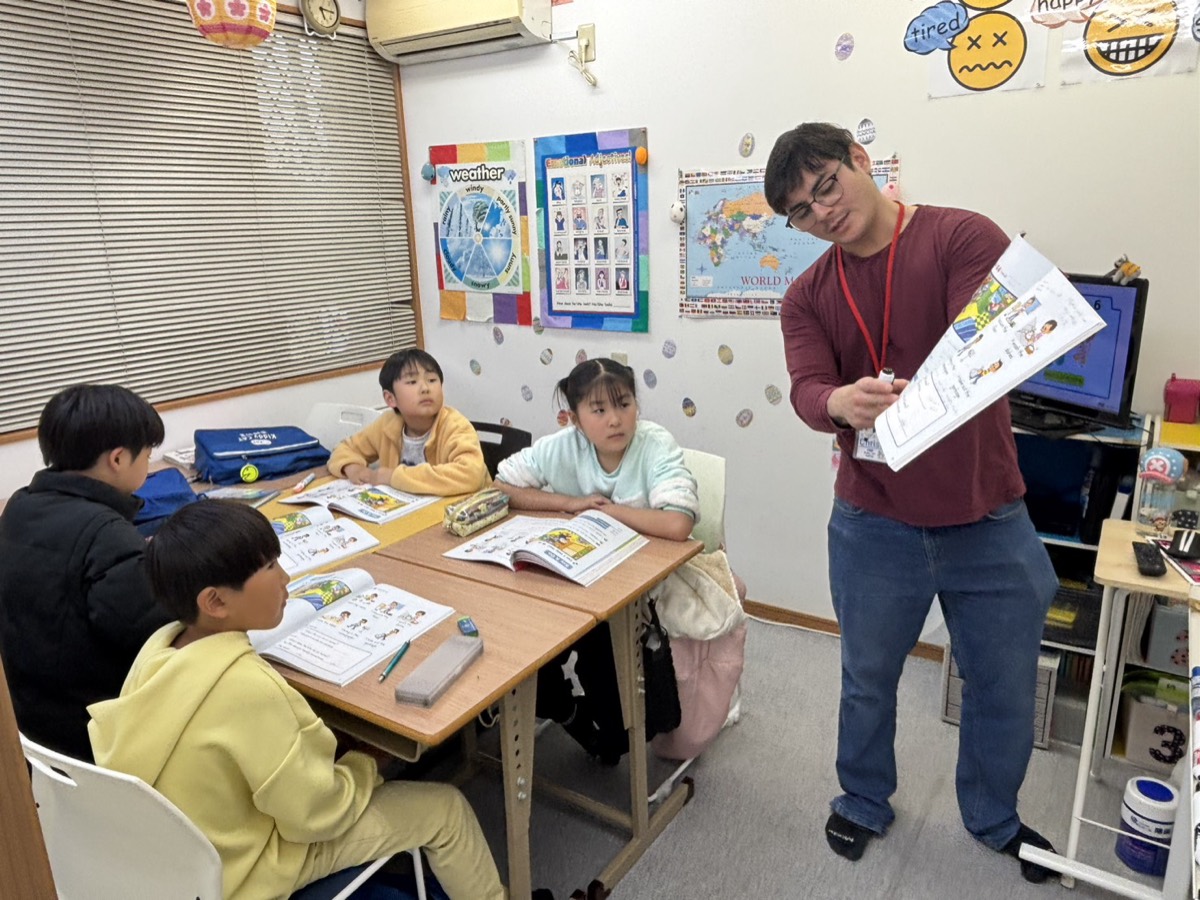 講師がテキストを見せながら、机でテキストを開く子どもたちにレッスンをしている様子