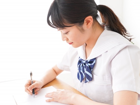 机に向かって、ペンでノートに書きこむ中学生